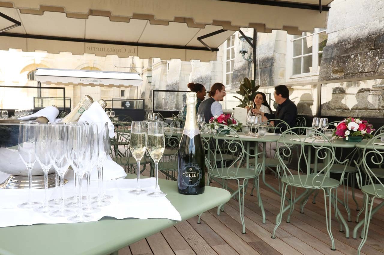 Terrasse d'un restaurant avec des tables en fer forgé, des verres et une bouteille de champagne au premier plan.
