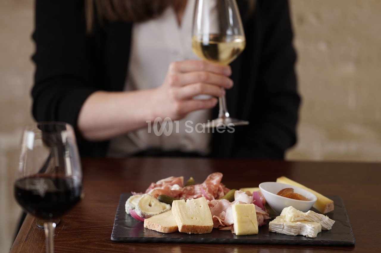 Plateau de fromages et charcuteries sur une table, avec une personne tenant un verre de vin blanc.