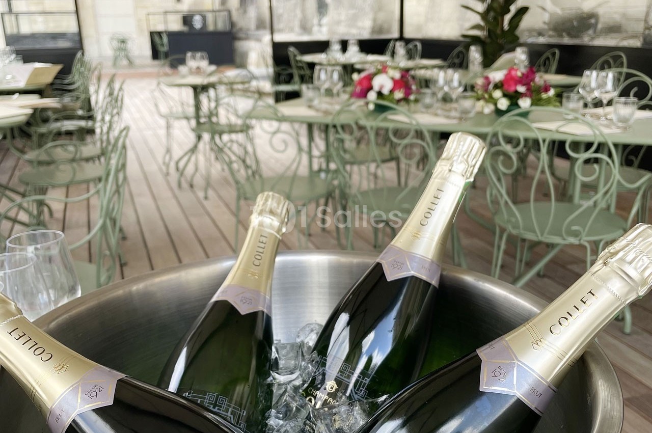 Bouteilles de champagne dans un seau à glace sur une terrasse avec tables et chaises en métal vert clair.