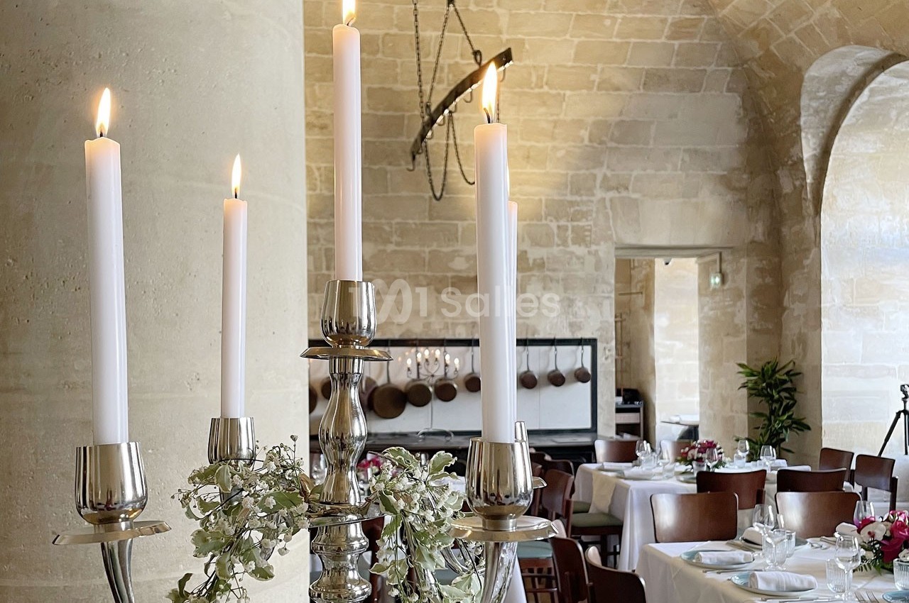 Chandelier argenté avec bougies allumées, décoré de feuillage, dans une salle de réception en pierre avec tables dressées.