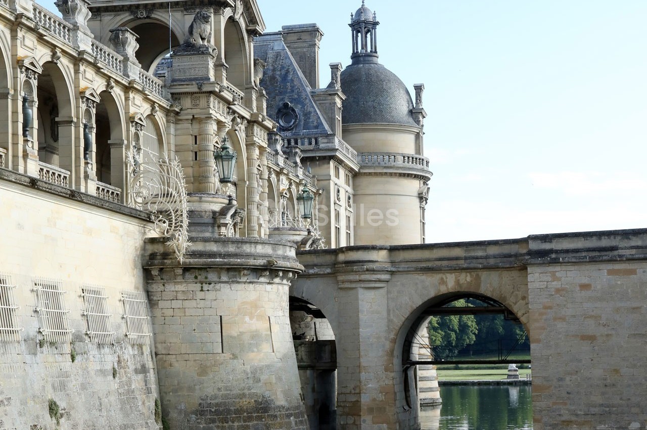 Façade d'un château en pierre avec arcades, tourelles et pont au-dessus d'un plan d'eau calme.