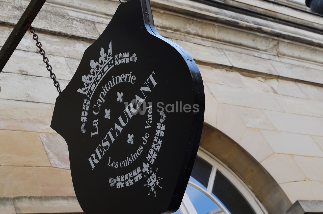 Enseigne noire suspendue indiquant ’La Capitainerie Restaurant, Les cuisines de Vatel’ devant un bâtiment en pierre.