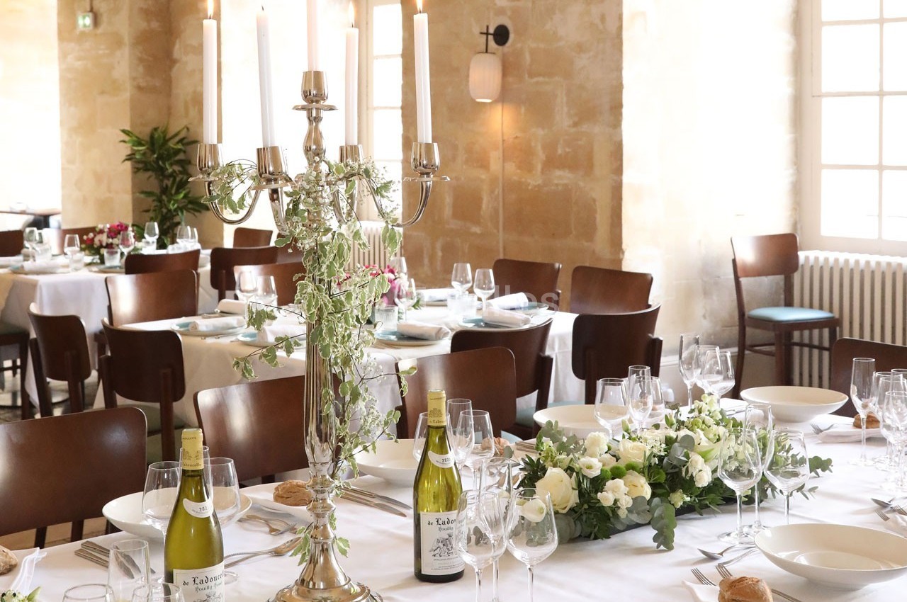 Salle de réception élégante avec tables dressées, chandeliers, fleurs blanches et bouteilles de vin.