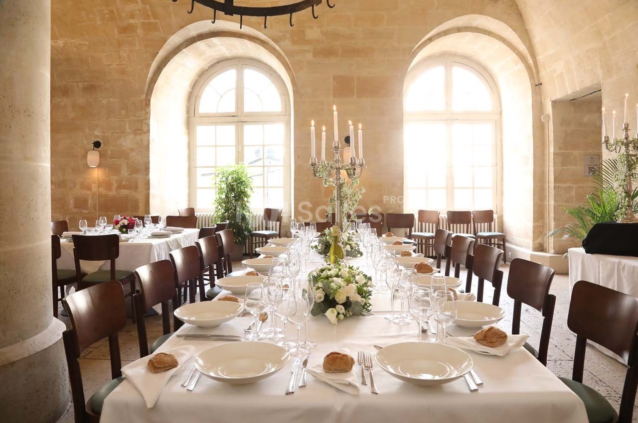 Salle lumineuse avec une grande table dressée pour un repas, ornée de fleurs, chandeliers et vaisselle élégante.