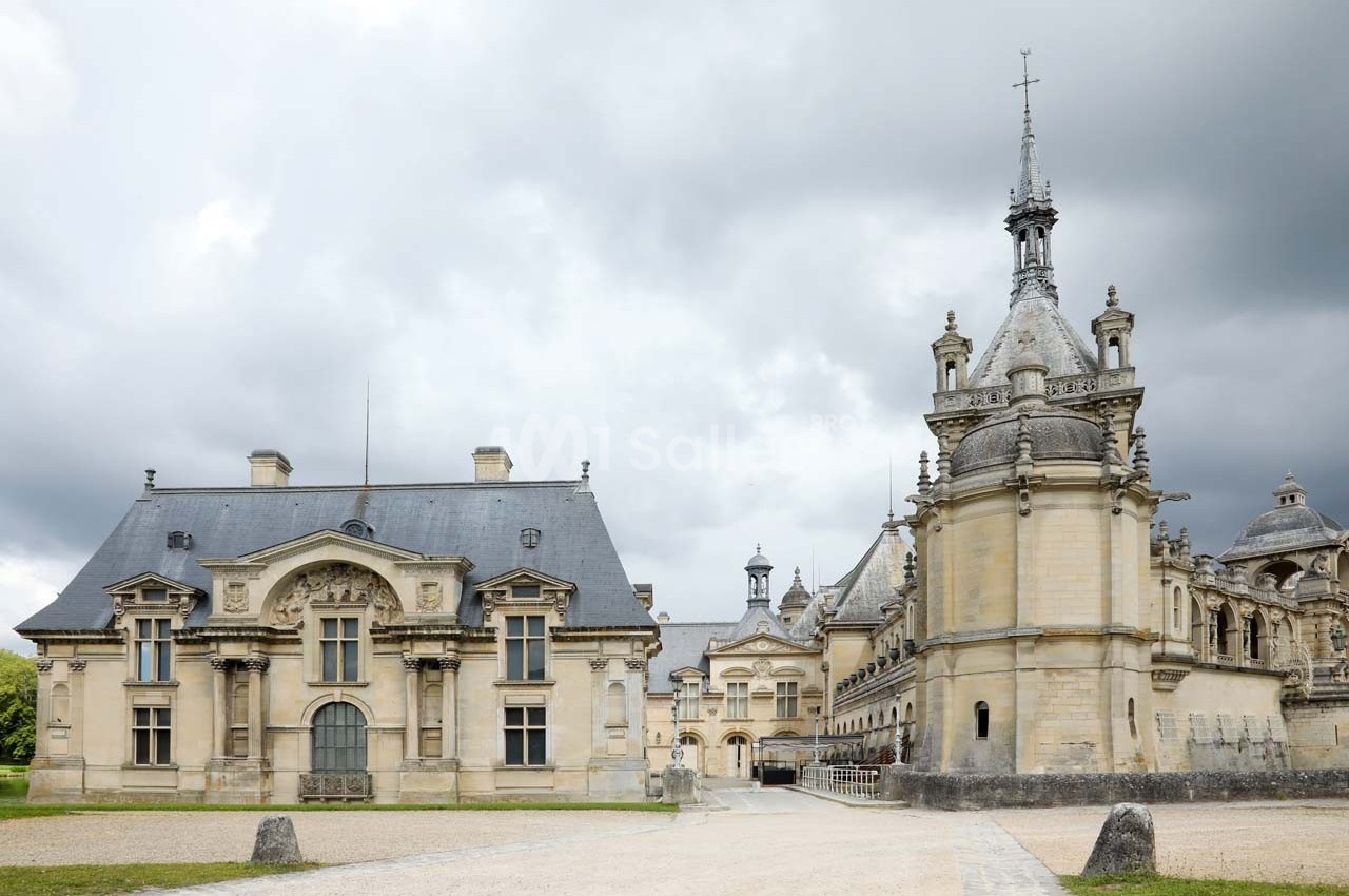 Façade d'un château historique avec des bâtiments en pierre, toits en ardoise et ciel nuageux en arrière-plan.