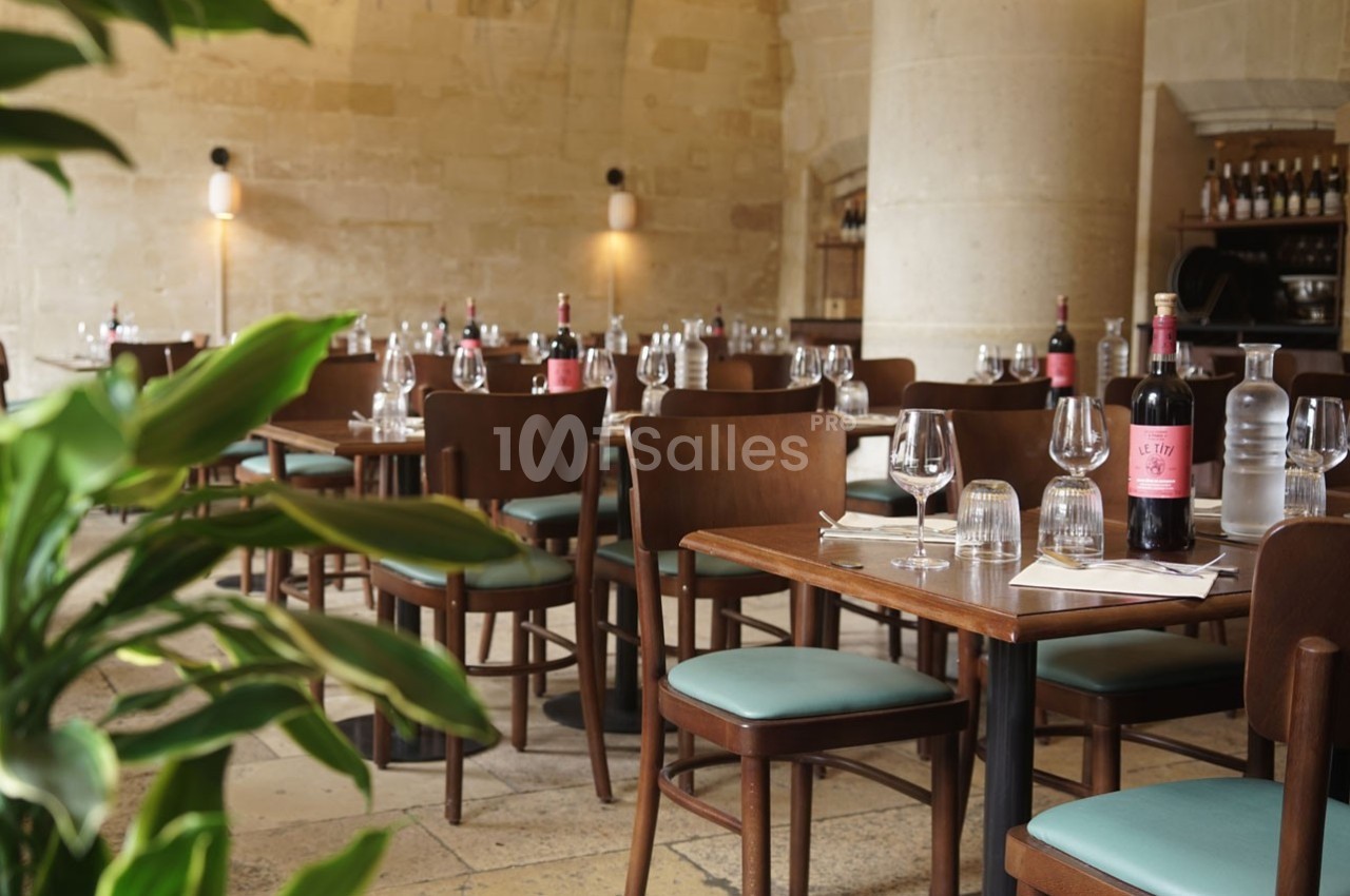 Salle de restaurant avec tables dressées, chaises en bois, bouteilles de vin et murs en pierre apparente.