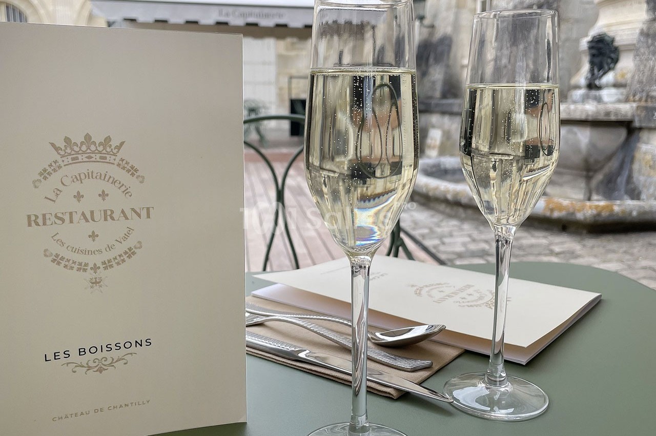 Deux verres de champagne sur une table de restaurant en terrasse, avec un menu et un décor en pierre en arrière-plan.
