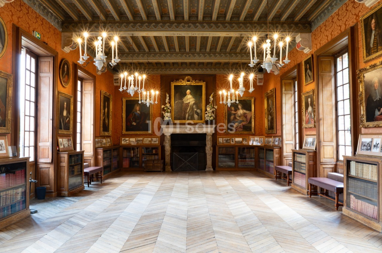 Salle ornée de portraits et de chandeliers, avec parquet en bois, bibliothèque et cheminée centrale.
