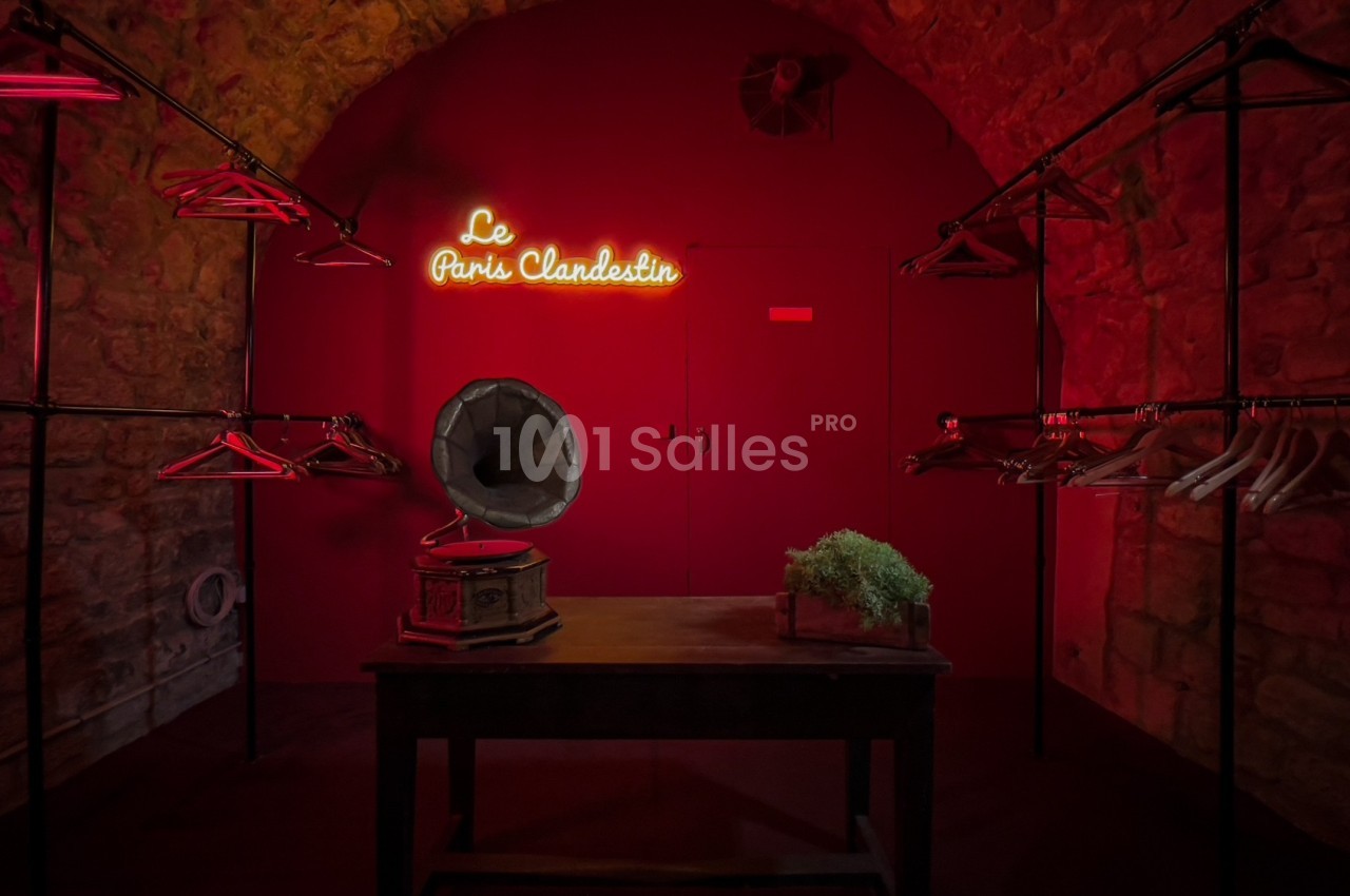 Salle en pierre éclairée de rouge avec un gramophone sur une table, des portants vides et une enseigne lumineuse.