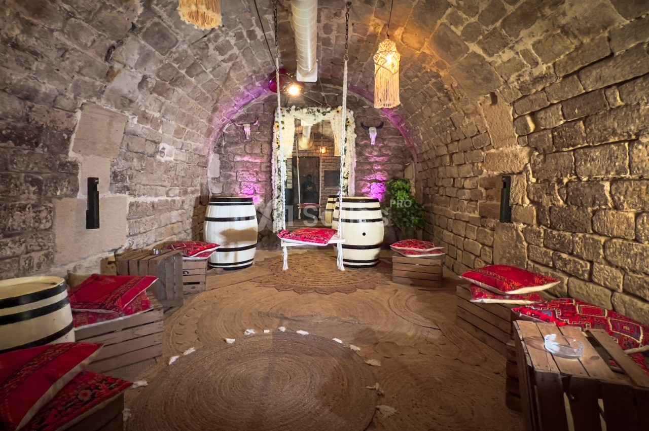 Salle voûtée en pierre avec des tonneaux, coussins colorés et éclairage tamisé, décorée dans un style rustique.