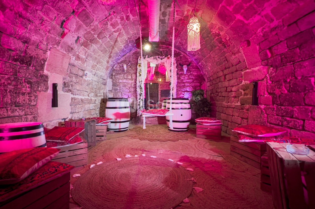 Salle voûtée en pierre avec éclairage rose, décorée de coussins, tonneaux et hamacs suspendus.