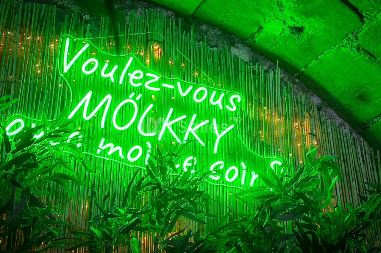 Enseigne néon verte ’Voulez-vous mölkky avec moi ce soir ?’ sur fond de bambou et feuillage.