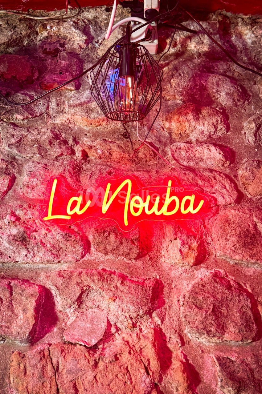 Enseigne néon rouge ’La Nouba’ accrochée à un mur en pierre, éclairée par une lampe suspendue.