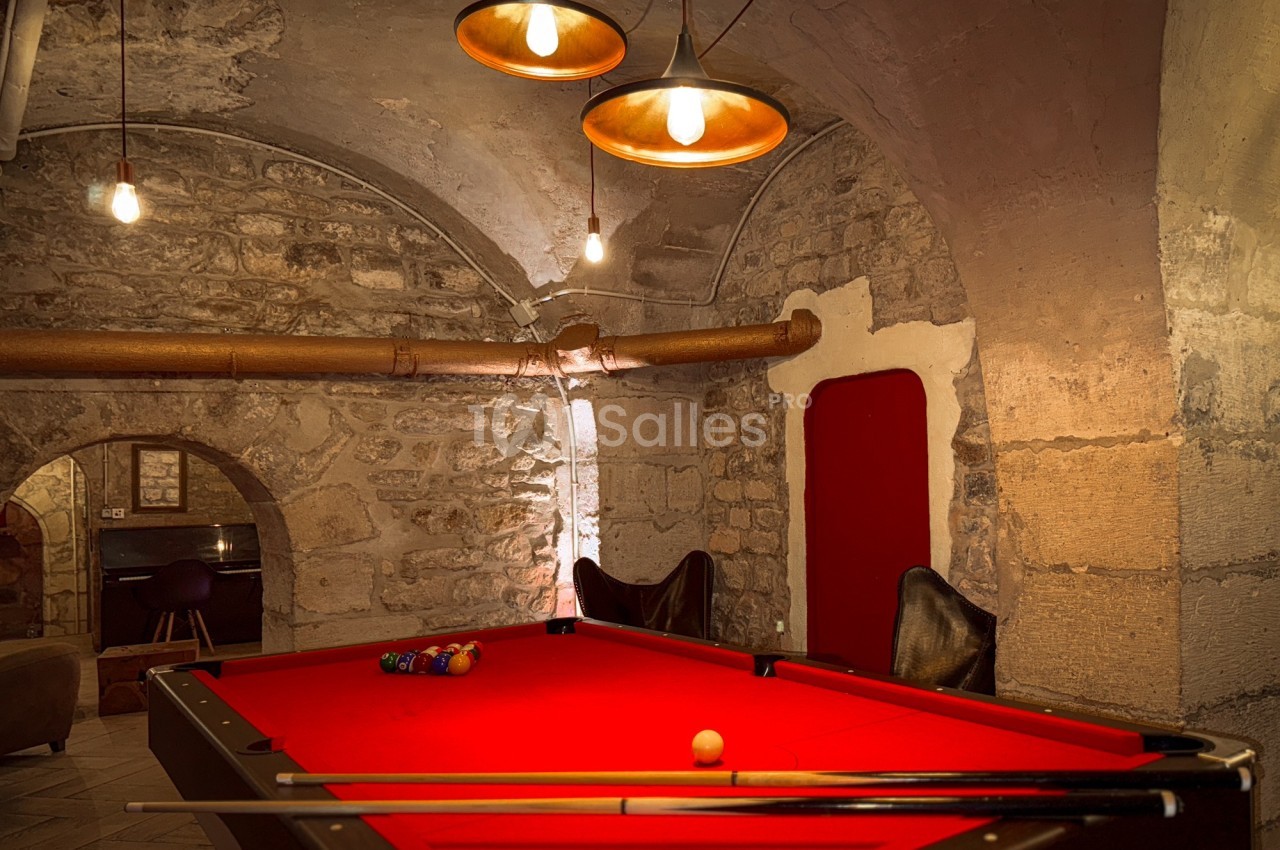 Table de billard rouge dans une pièce voûtée en pierre, éclairée par des suspensions lumineuses.
