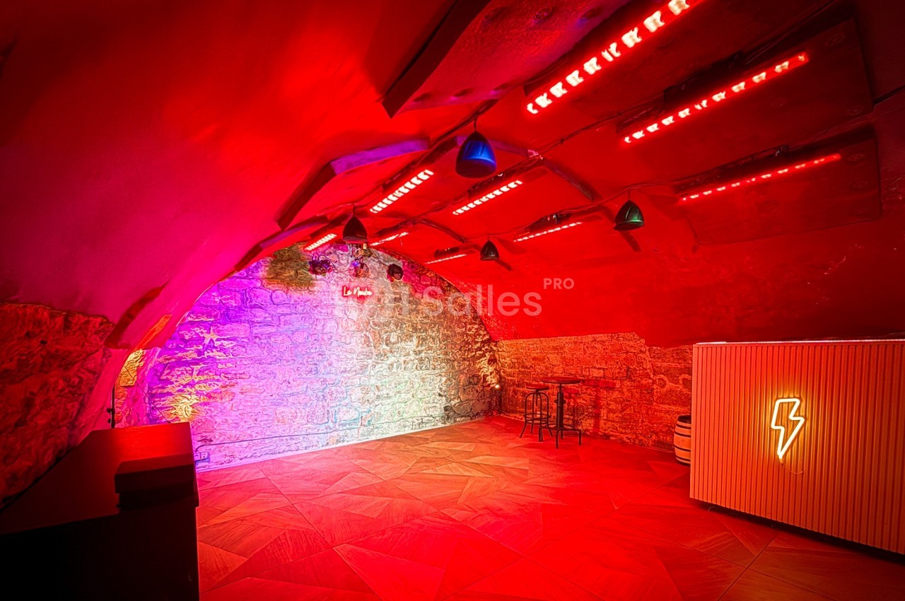 Salle voûtée en pierre éclairée par des lumières rouges et colorées, avec un bar et quelques tabourets.