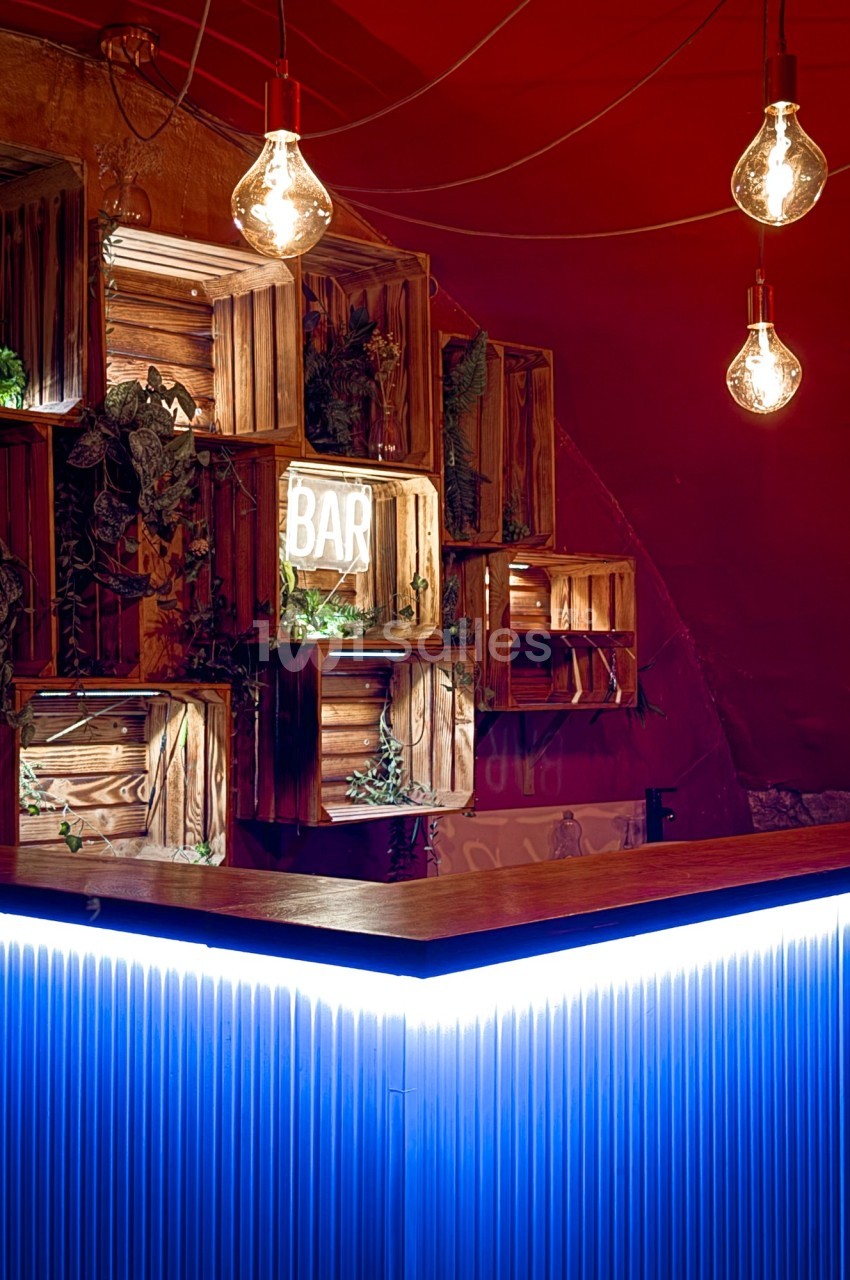 Comptoir de bar éclairé par des néons bleus, entouré de caisses en bois décorées de plantes et d'ampoules suspendues.