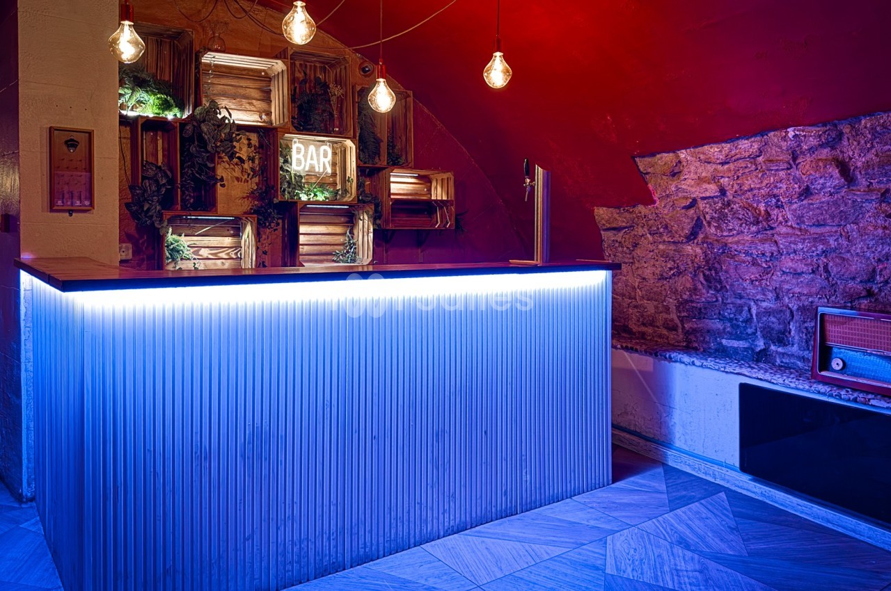Comptoir de bar rétro éclairé en bleu, avec des étagères décoratives, des ampoules suspendues et un mur en pierre.