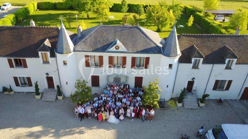 Vue aérienne d'un groupe de personnes rassemblées devant un grand bâtiment de style manoir entouré de verdure.
