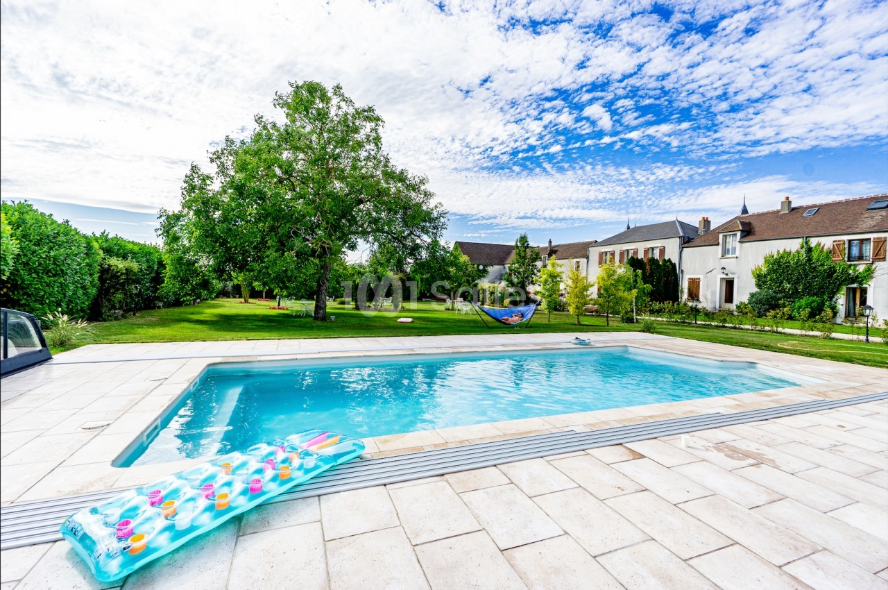 Piscine extérieure entourée d'une terrasse en pierre, avec jardin arboré et maisons en arrière-plan sous un ciel bleu.