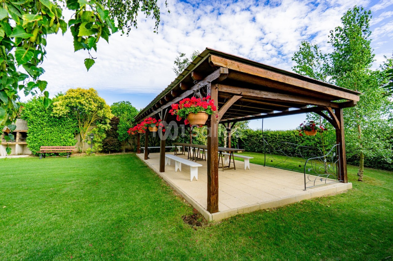 Pergola en bois avec tables et bancs, entourée de pelouse, fleurs suspendues et arbres dans un jardin.