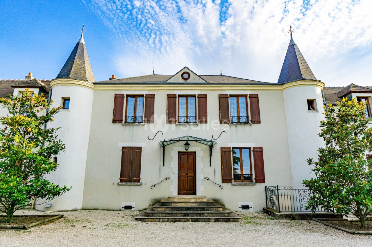 Façade d'un petit château blanc avec des volets en bois, des tours aux extrémités et un ciel partiellement nuageux.