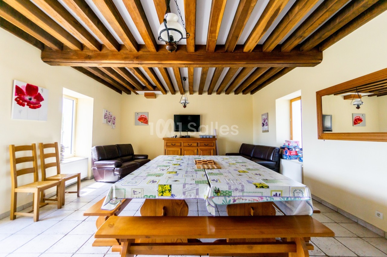 Salle à manger lumineuse avec poutres apparentes, grande table en bois, chaises, canapé et télévision au mur.