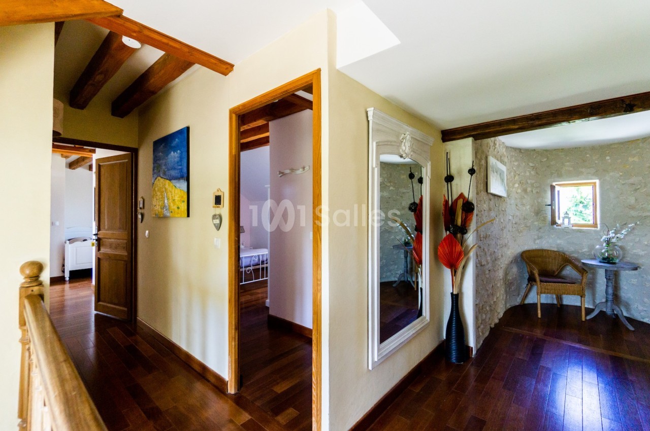 Vue d'un couloir intérieur avec parquet, poutres apparentes, miroir mural et décoration florale près d'une fenêtre.
