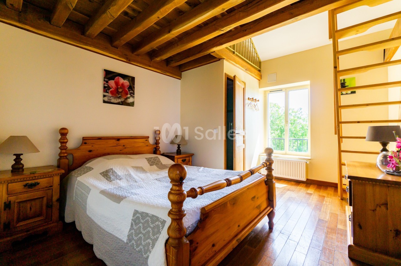 Chambre lumineuse avec lit en bois, plafond en poutres apparentes, échelle menant à une mezzanine et fenêtre donnant sur la…