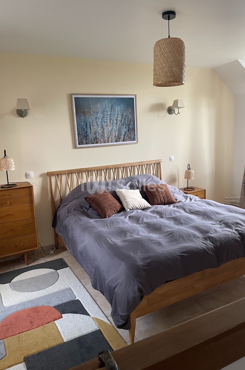 Chambre lumineuse avec lit double, linge gris, coussins décoratifs, mobilier en bois et tableau mural.