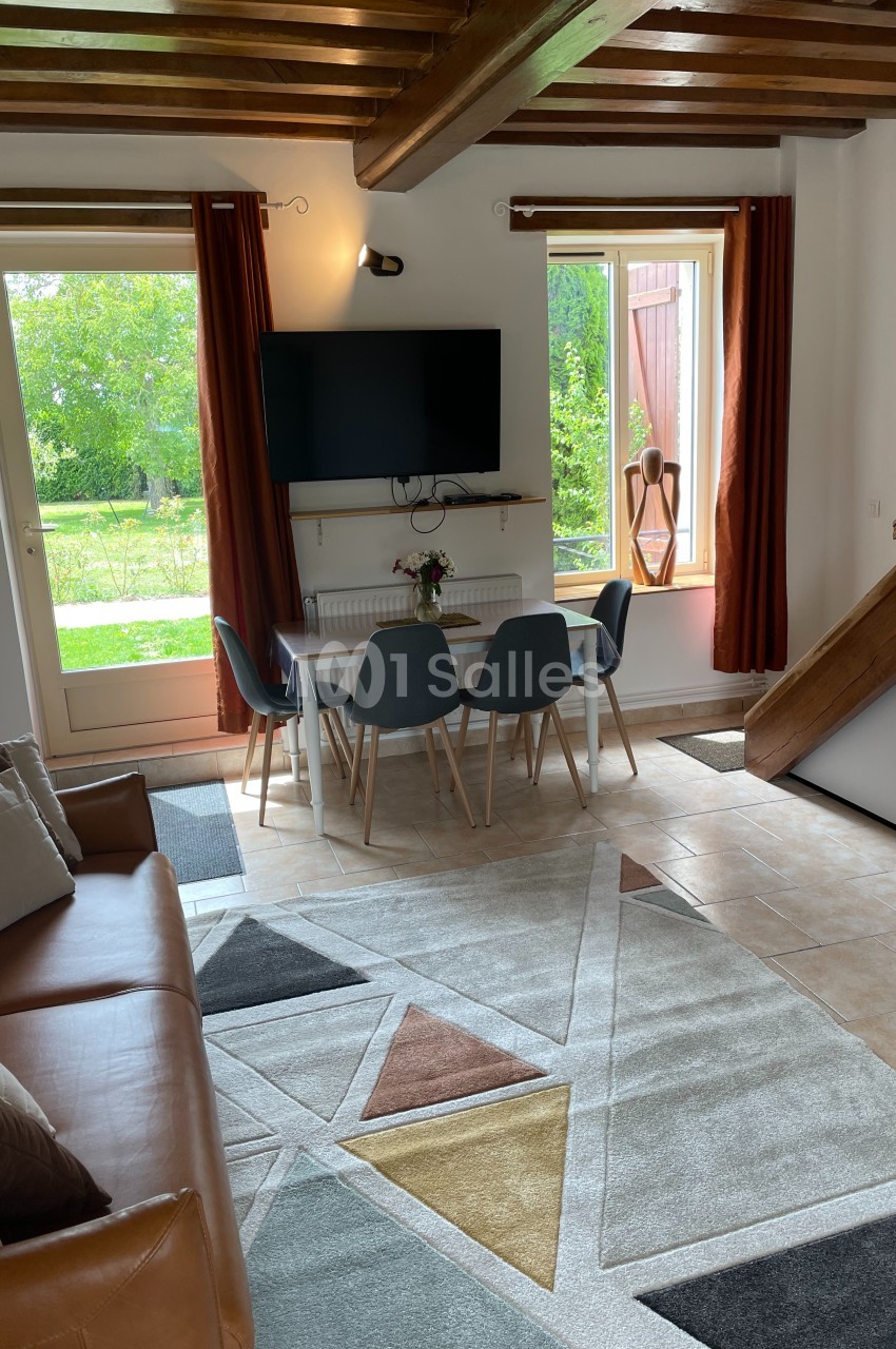Salon lumineux avec canapé, table à manger, quatre chaises, télévision murale, tapis géométrique et vue sur jardin.