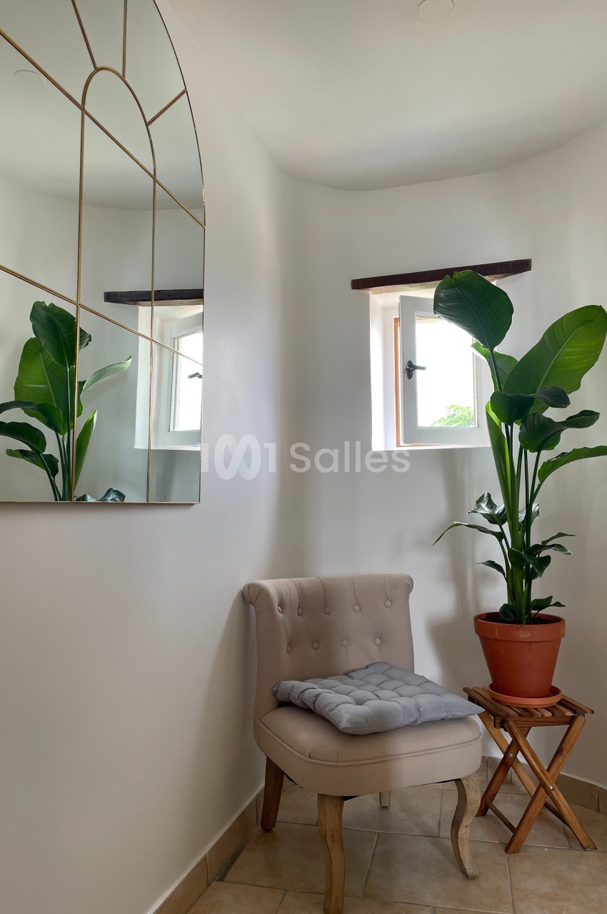 Fauteuil beige avec coussin, plante en pot sur un tabouret et miroir doré dans un coin lumineux avec deux petites fenêtres.