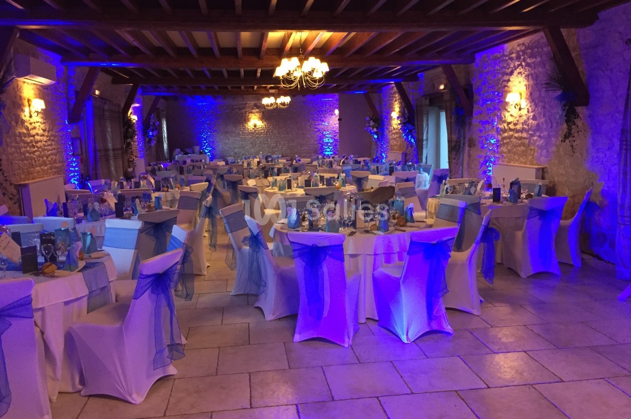 Salle de réception décorée pour un événement, avec des tables rondes, nappes blanches et éclairage bleu.