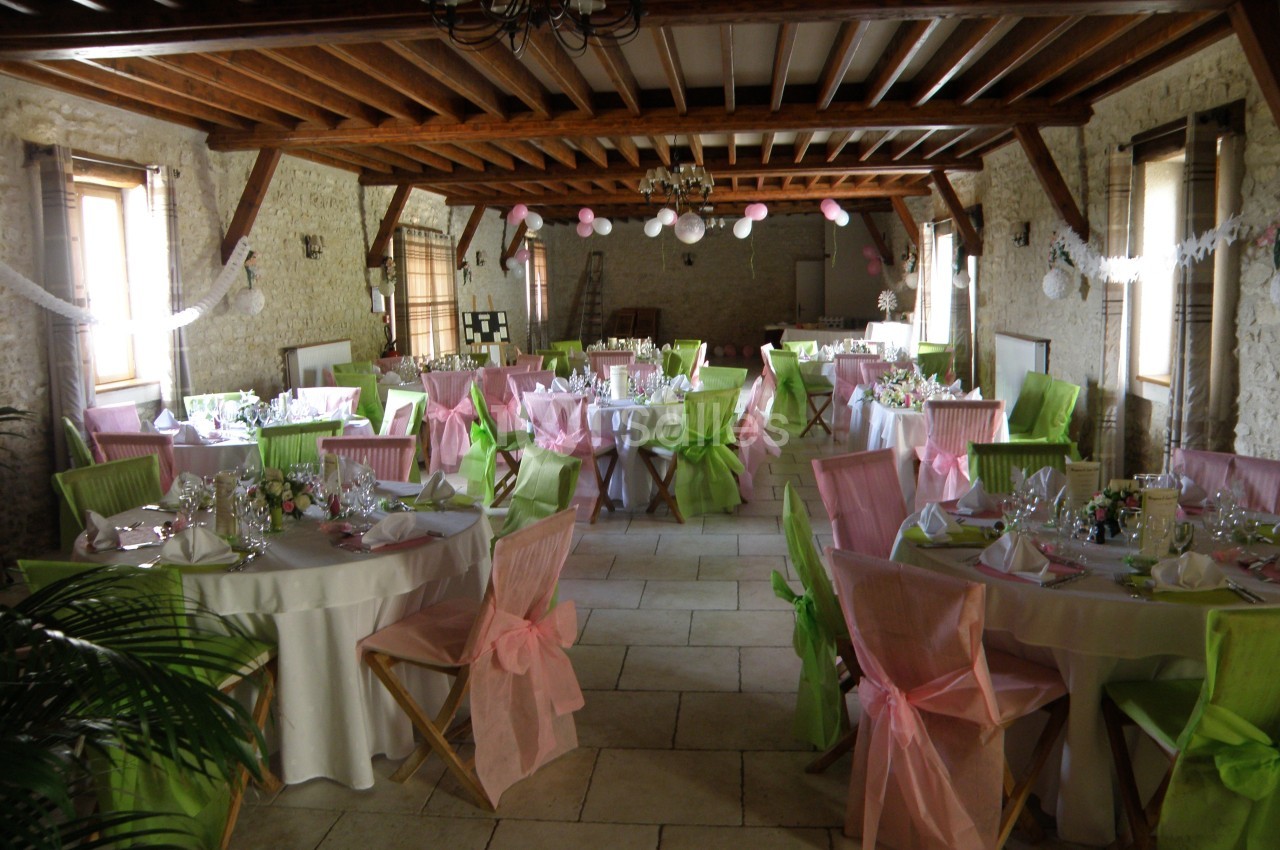 Salle de réception décorée avec des tables rondes, nappes blanches et chaises ornées de nœuds roses et verts.