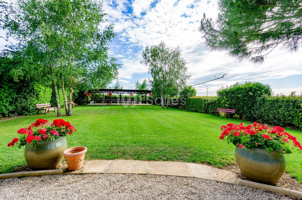 Jardin verdoyant avec pelouse, pots de fleurs rouges, pergola ombragée et bancs en bois sous un ciel partiellement nuageux.
