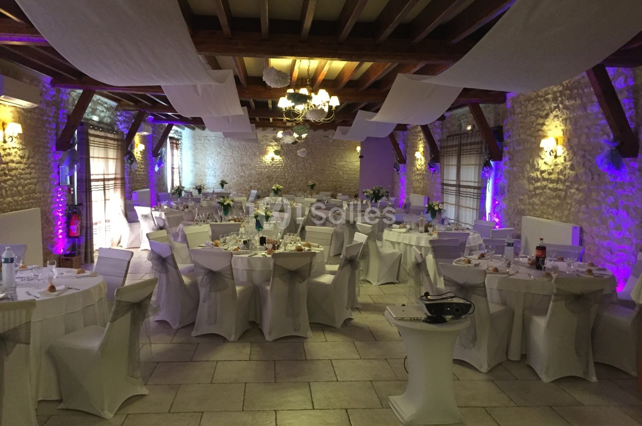 Salle de réception décorée avec des tables rondes nappées de blanc, éclairage tamisé et poutres apparentes.