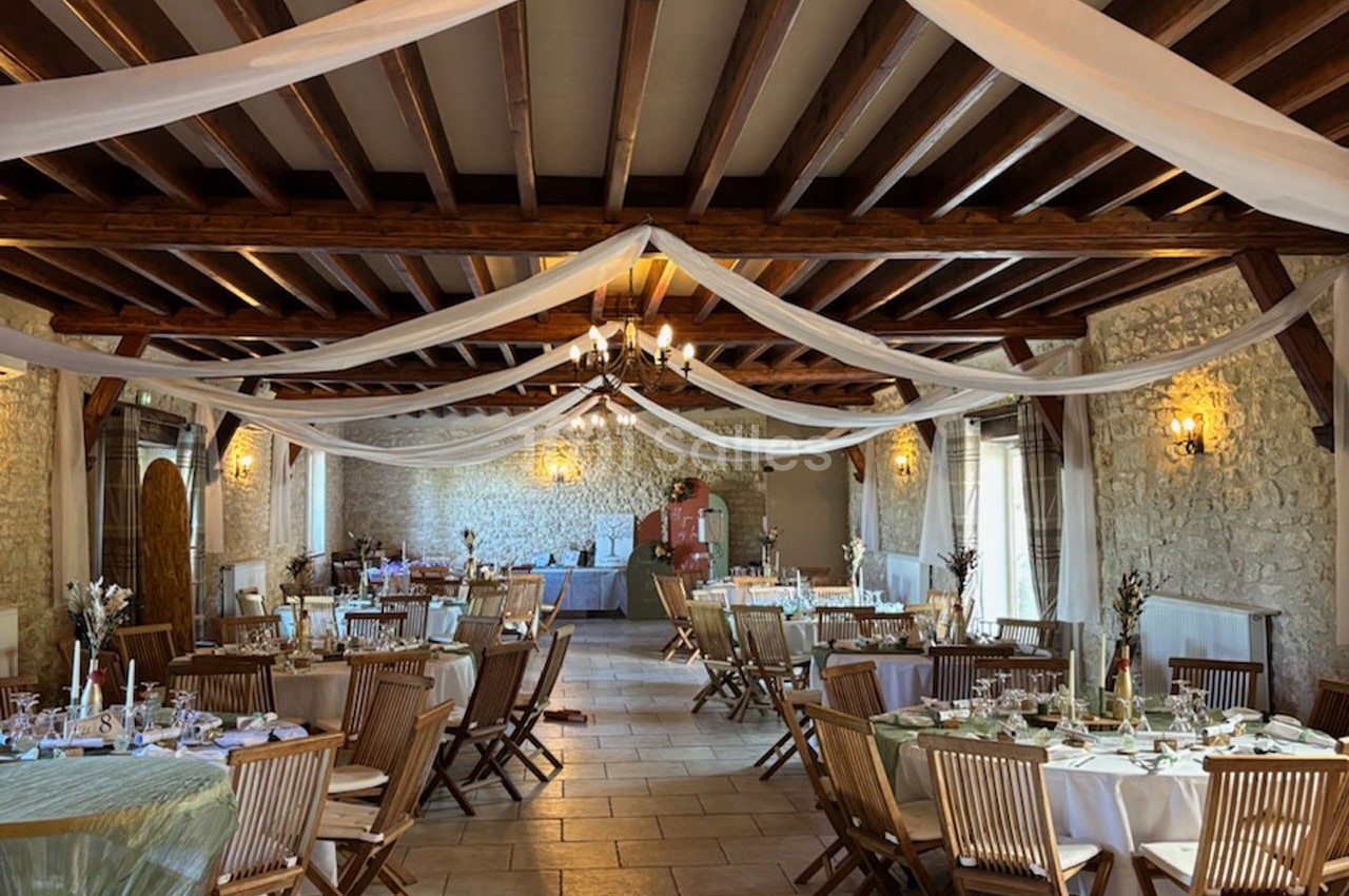 Salle de réception décorée avec des tables rondes, nappes blanches, chaises en bois et drapés suspendus au plafond.
