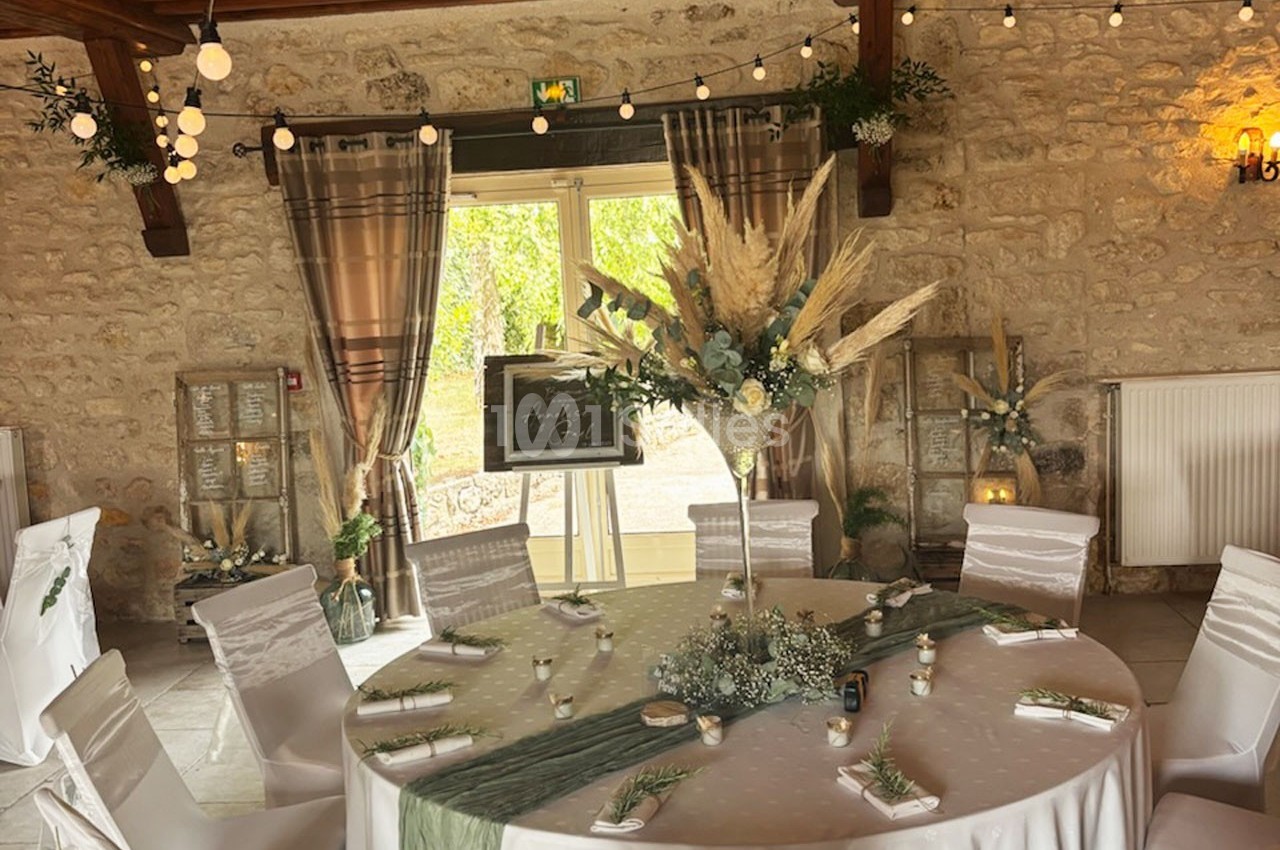 Salle de réception décorée avec une table ronde ornée de fleurs séchées, nappes blanches et guirlandes lumineuses.