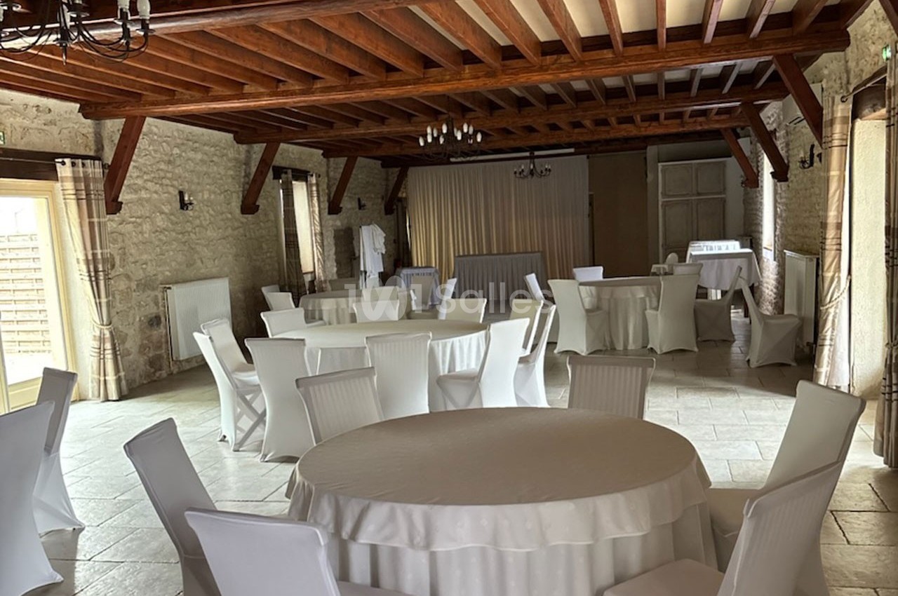 Salle de réception en pierre avec tables rondes et chaises blanches, éclairée par la lumière naturelle des fenêtres.