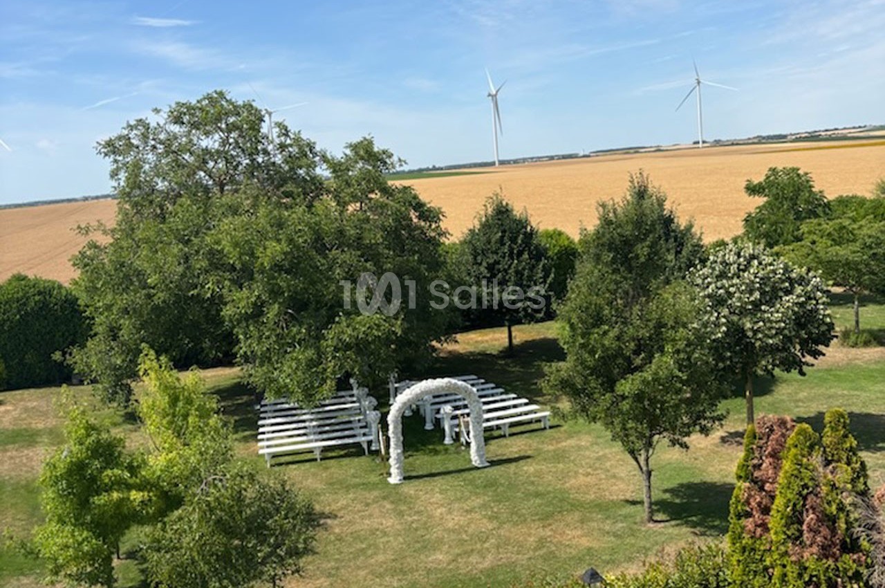 Vue d'un jardin avec des bancs blancs disposés devant une arche décorée, entouré d'arbres et de champs avec des éoliennes au…