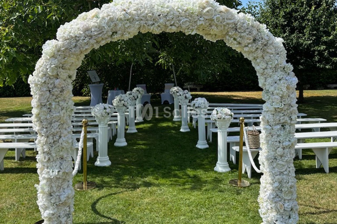 Arche de fleurs blanches menant à une allée bordée de bancs et de colonnes fleuries dans un jardin verdoyant.