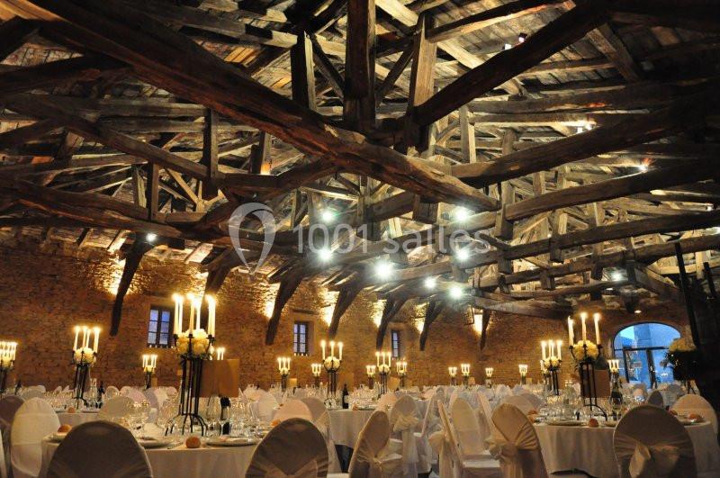 Salle de réception avec charpente en bois apparente, tables dressées et chandeliers allumés.