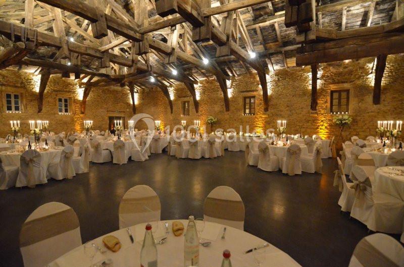 Salle de réception en pierre avec charpente apparente, tables rondes dressées et chandeliers allumés.