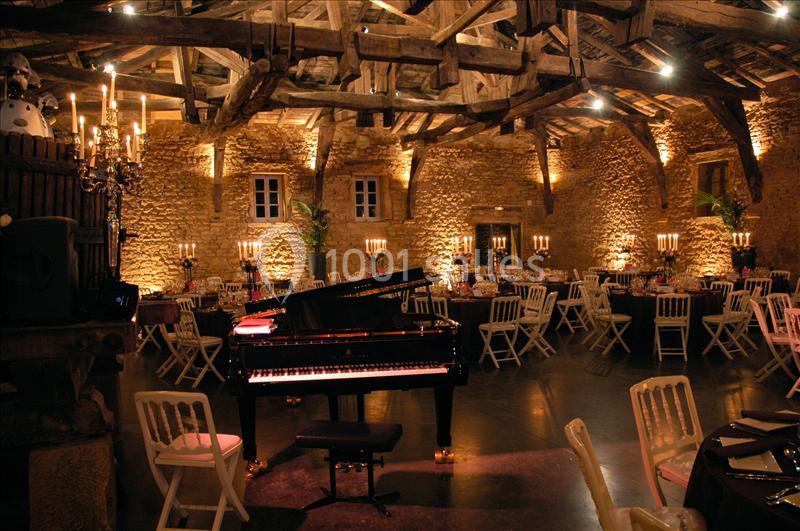 Salle en pierre avec charpente apparente, éclairée par des chandeliers, tables dressées et un piano à queue au centre.