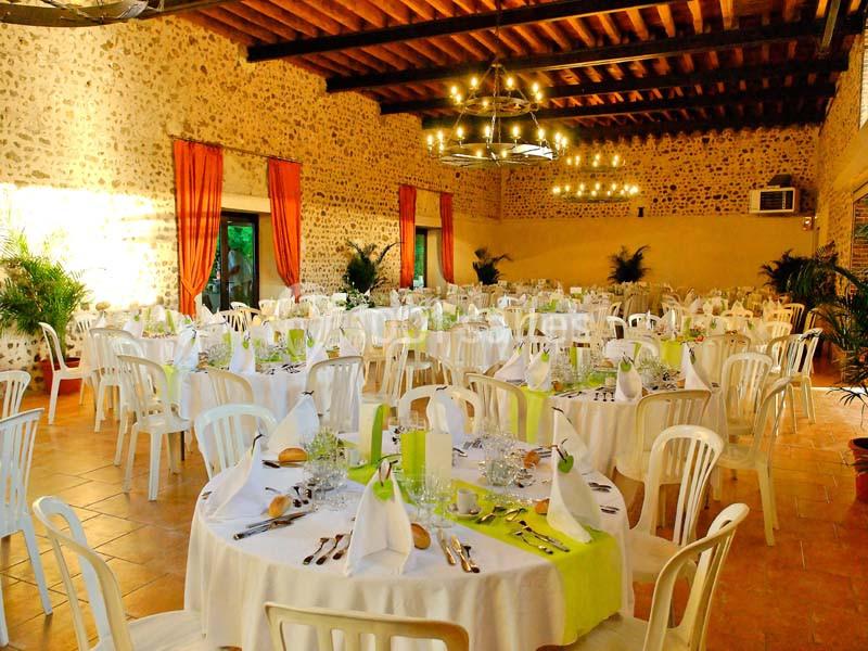 Salle de réception lumineuse avec tables rondes dressées, nappes blanches et décorations vertes, murs en pierre apparente.