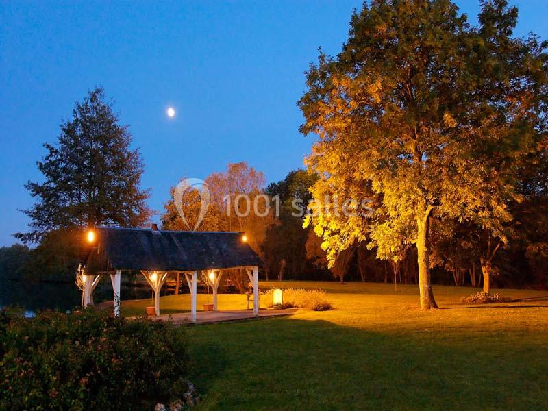 Abri en bois éclairé sous un ciel nocturne, entouré d'arbres et d'une pelouse.