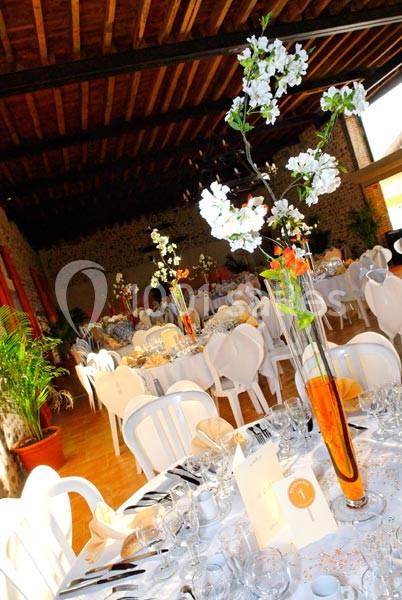 Salle de réception décorée avec des tables rondes dressées, centres de table fleuris et chaises blanches.