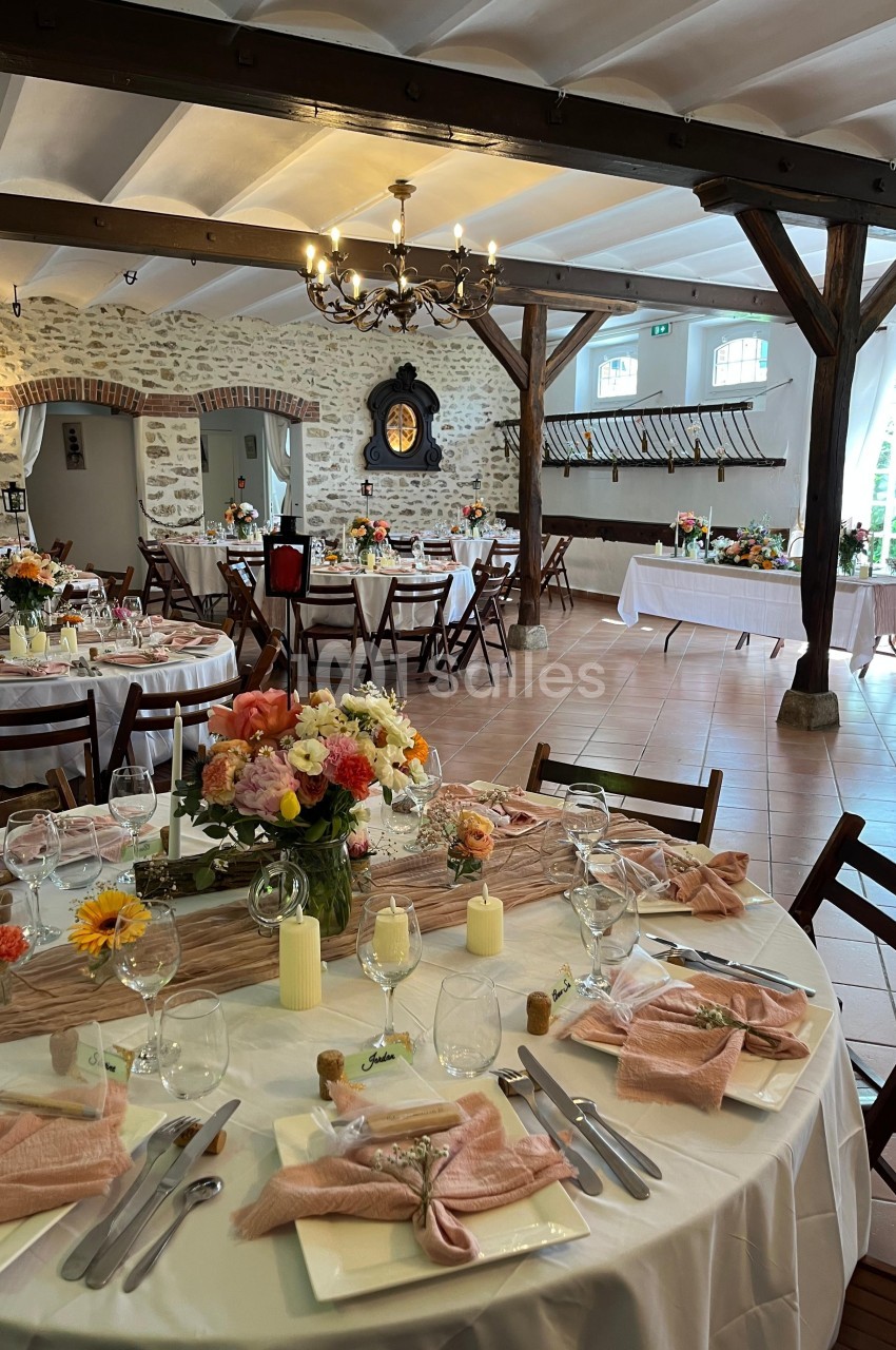 Salle de réception décorée pour un événement, avec tables dressées, nappes blanches, fleurs et lumière naturelle.