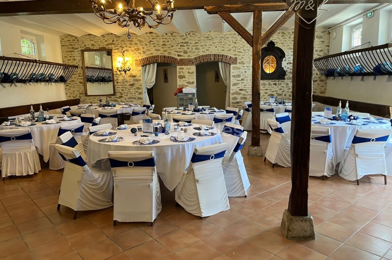 Salle de réception décorée avec des tables rondes dressées, nappes blanches et chaises ornées de rubans bleus.