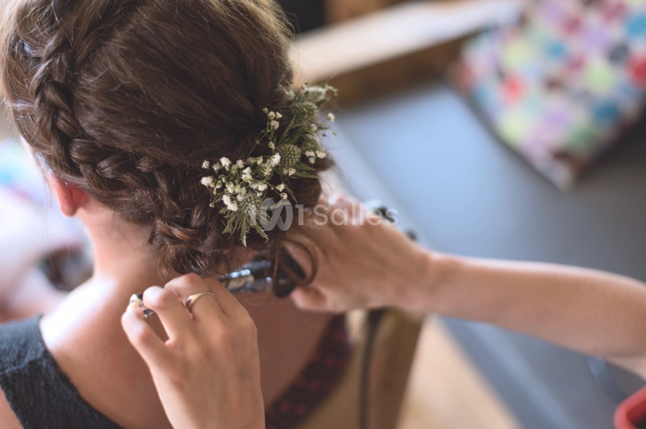 Une femme aux cheveux tressés ornés de fleurs blanches se fait coiffer dans un cadre intérieur lumineux.