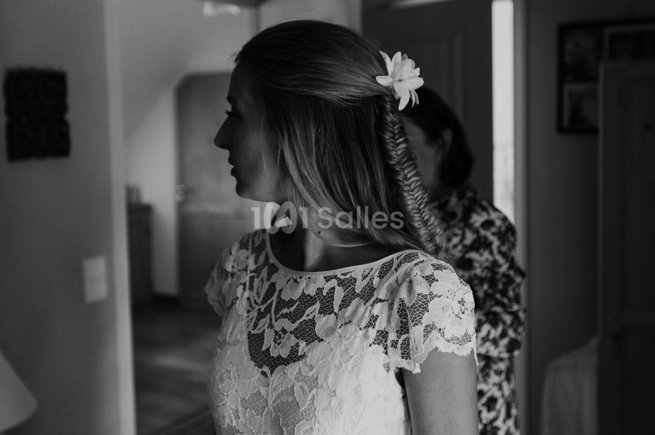 Femme en robe en dentelle blanche, coiffée d'une tresse et d'une fleur, dans un intérieur lumineux.